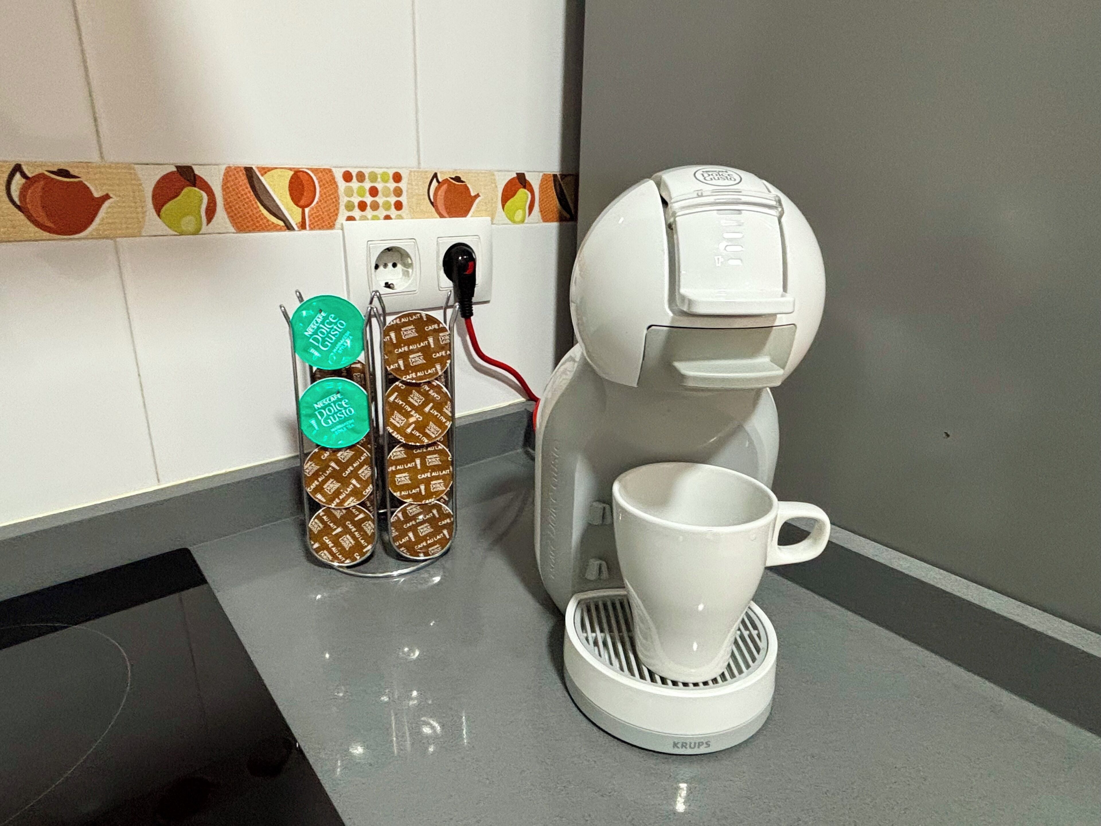 A coffee maker with a white mug on a grey countertop, a power outlet, and a wall with a fruit pattern backsplash.