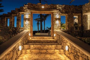 Una escalera de piedra conduce a una puerta de madera enmarcada por paredes de piedra, con luces a lo largo de los escalones y palmeras al fondo.