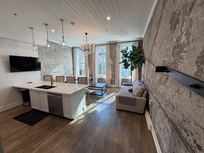 A modern kitchen with a central island, dining area with chairs, a sofa, and a large window with curtains. - Jimmys on Canal St  (New Orleans)