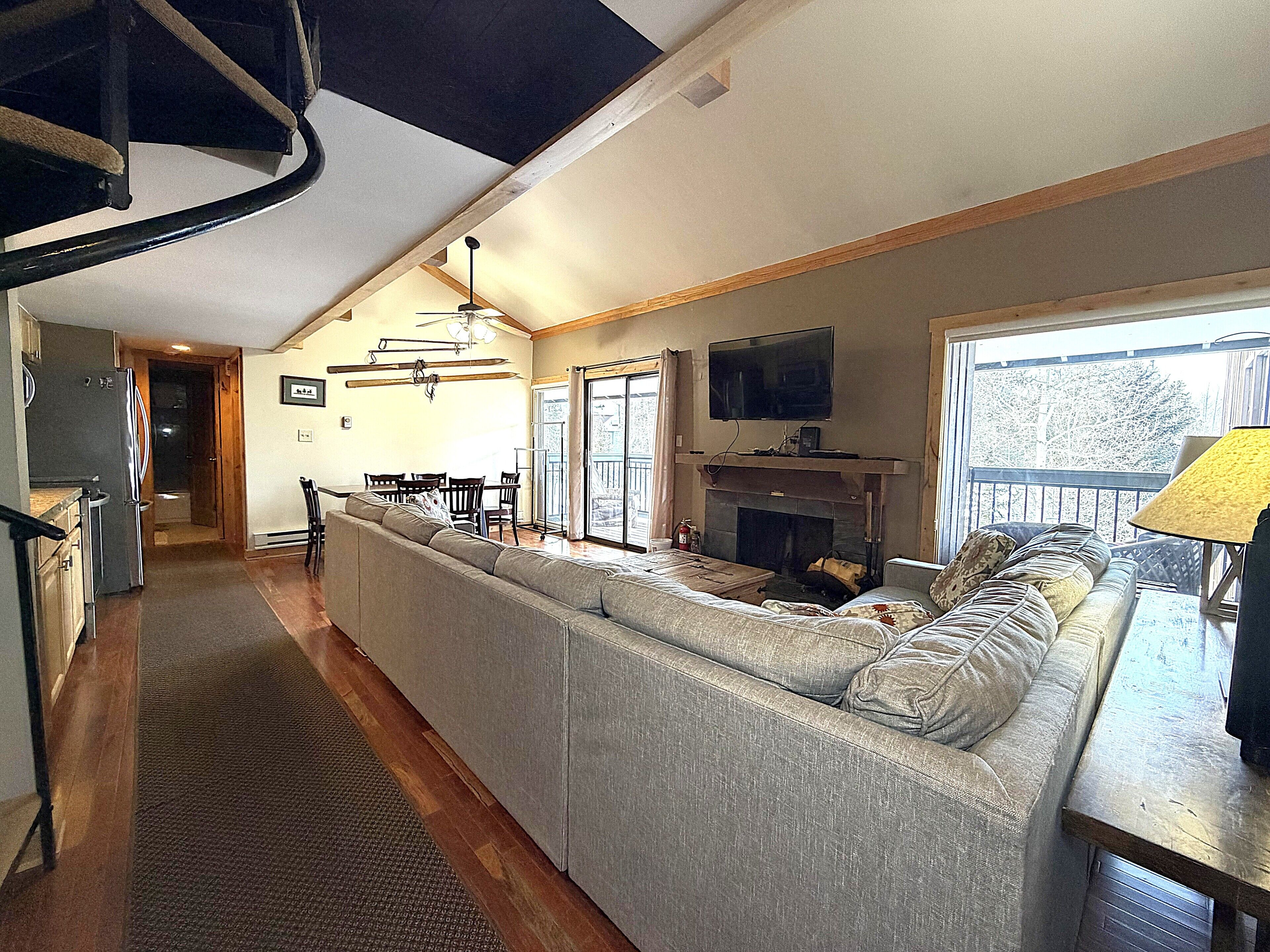 A spacious living area with a large sofa, a television mounted above a fireplace, and a dining area with a table and chairs.