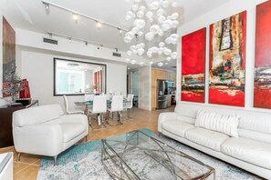 A modern living room with a glass coffee table, white sofas, a dining area with a white table and chairs, and abstract red paintings on the wall. - Sapphire 601 - By GISO Vacation Rentals (South Padre Island)