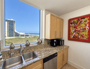 A kitchen with granite countertops, stainless steel appliances, wooden cabinets, and a large window with a view of buildings and the ocean. - Sapphire 601 - By GISO Vacation Rentals (South Padre Island)