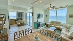 Amplia sala de estar con mesa de comedor de madera, un conjunto de sofás y una cocina con un amplio ventanal con vista al océano.