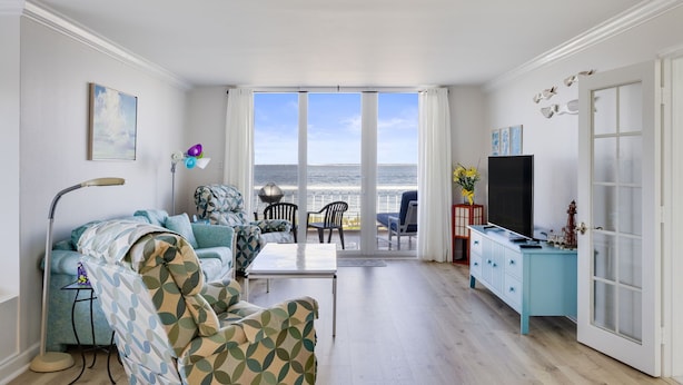 Een woonkamer met een bank, stoelen, een salontafel en een televisie. Er is een balkon met uitzicht op de zee.