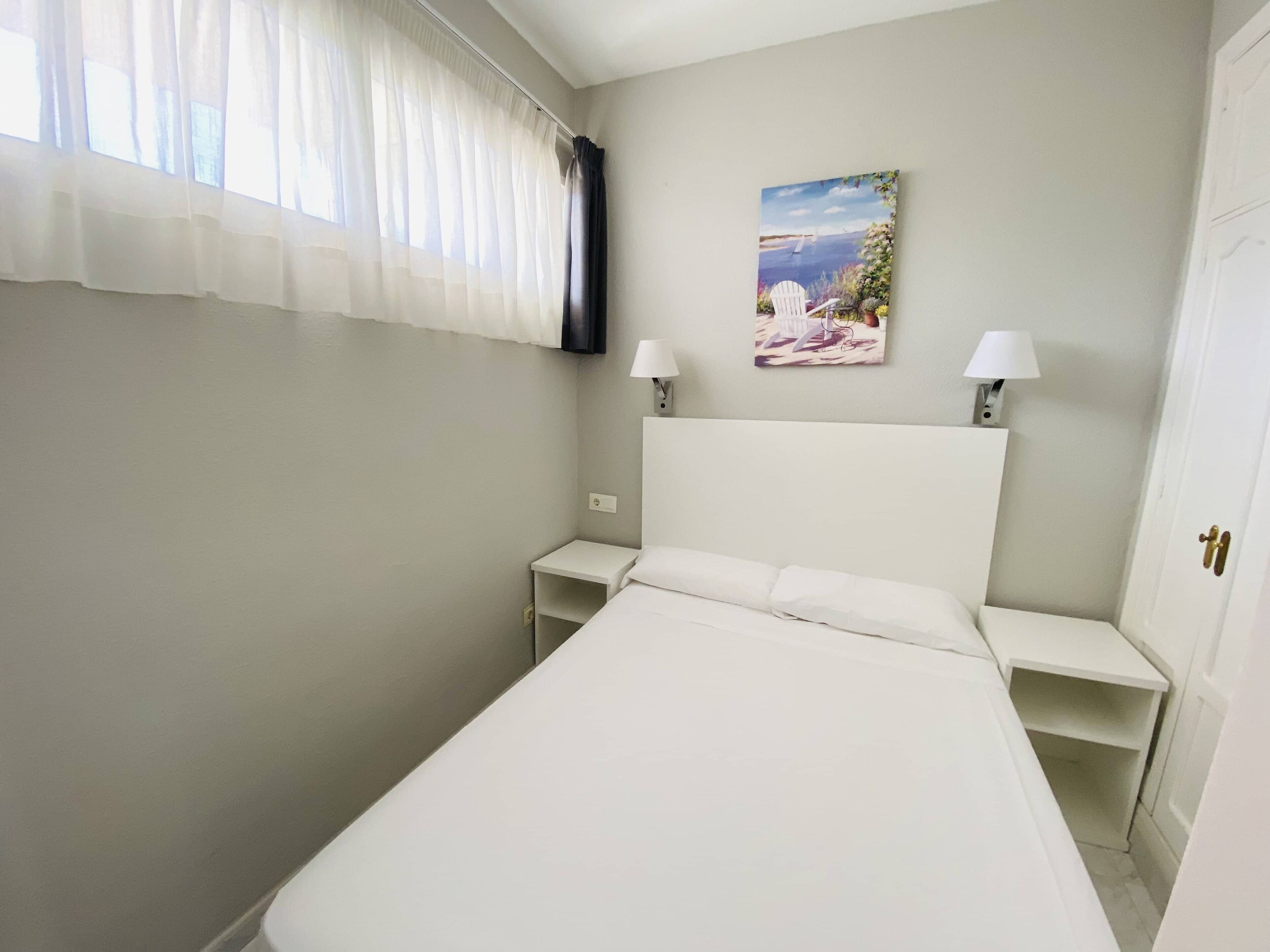 Una camera da letto con un letto, due comodini, un tavolino, un quadro e una finestra con tende leggere.