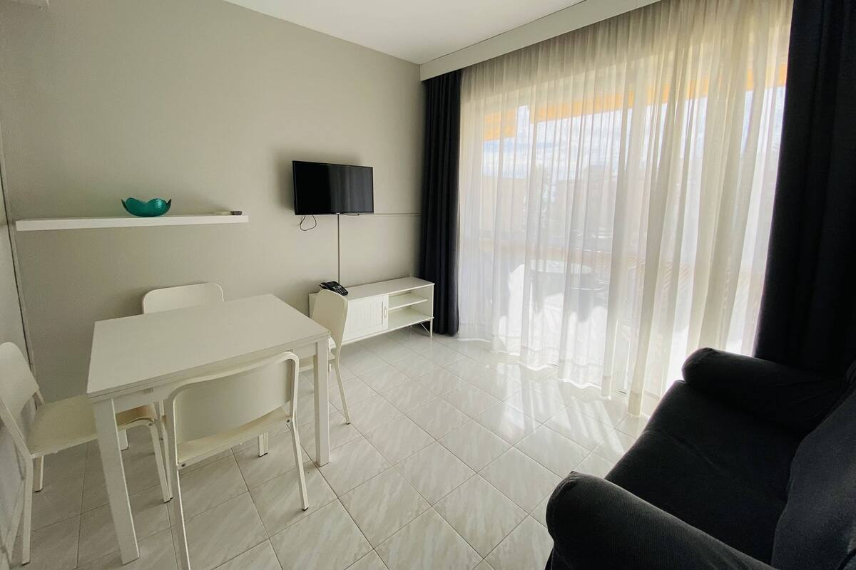 A modern living room with a white sofa, a dining table with chairs, a flat-screen TV mounted on the wall, and a large window with sheer curtains.