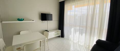 A modern living room with a white sofa, a dining table with chairs, a flat-screen TV mounted on the wall, and a large window with sheer curtains.