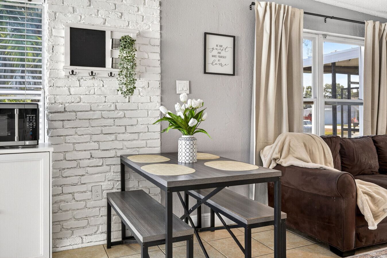 A modern living room with a white brick wall, a dining table with two chairs, a brown leather sofa, and a window with curtains.