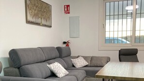 Una sala de estar moderna con un sofá seccional gris, dos sillones, una mesita y un extintor, además de un ventanal con vistas al exterior.