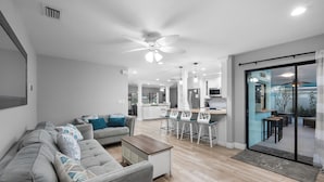 Ein modernes Wohnzimmer mit einem grauen Ecksofa, einem Couchtisch und einem Essbereich mit weißen Stühlen sowie einer Glastür zum Balkon.