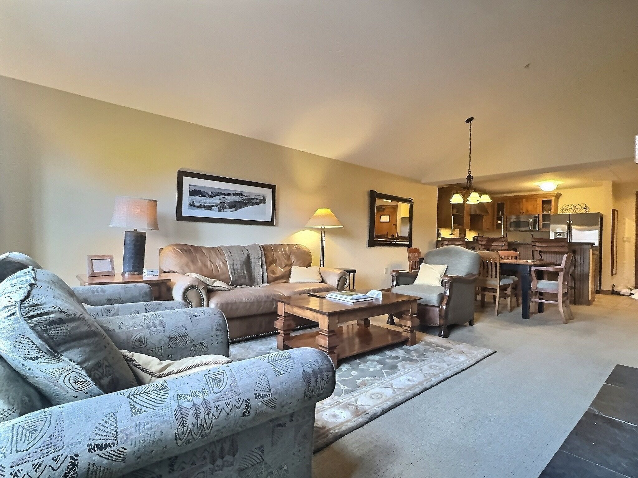 A living room with a sofa, armchairs, a coffee table, and a dining area with chairs and a table. There is a kitchen in the background with a refrigerator and cabinets.