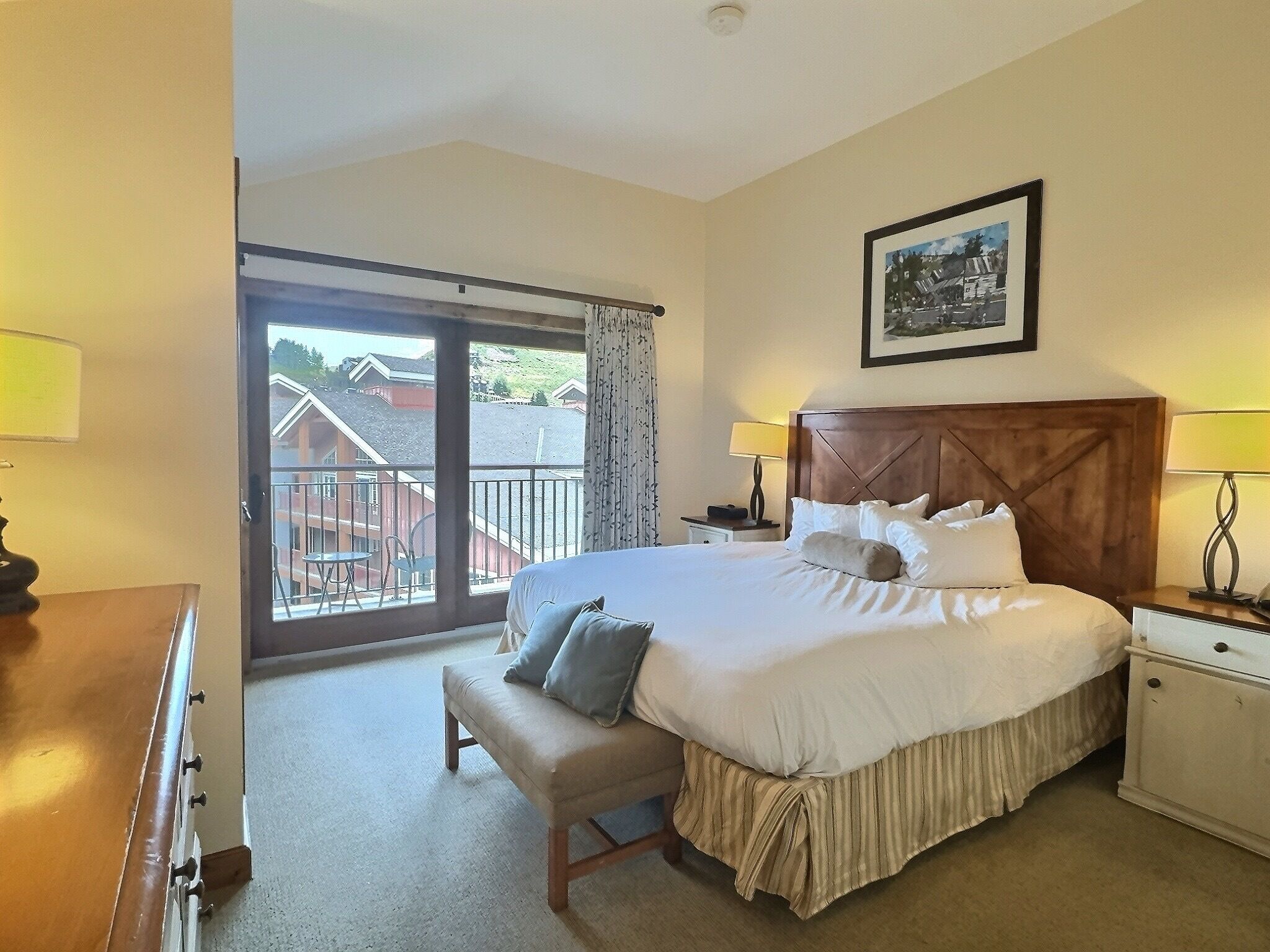 A hotel room with a large bed, a bench, a nightstand, a lamp, a framed picture on the wall, and a balcony with a view of buildings.