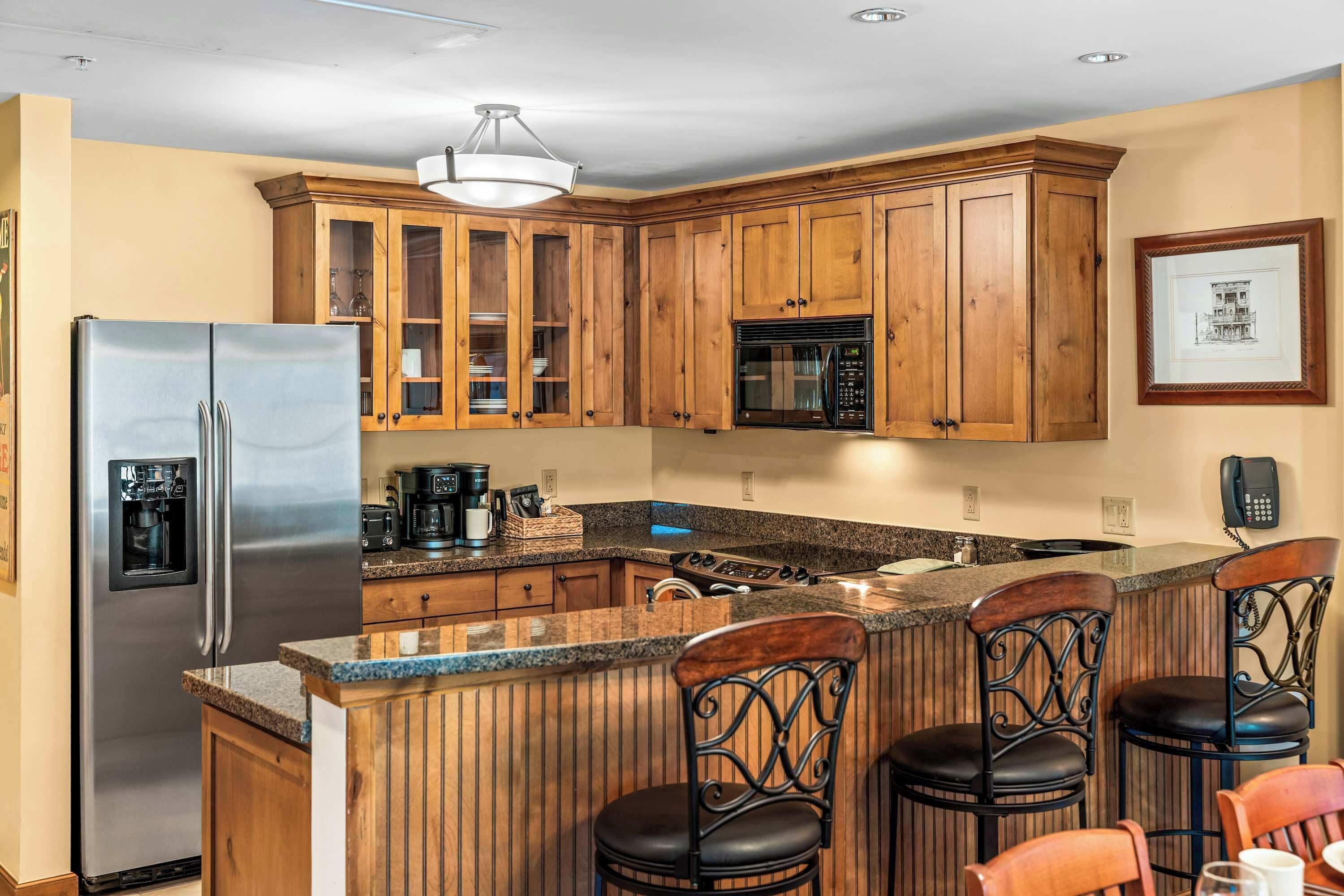 A kitchen with wooden cabinets, a stainless steel refrigerator, a built-in oven, and a bar area with stools.