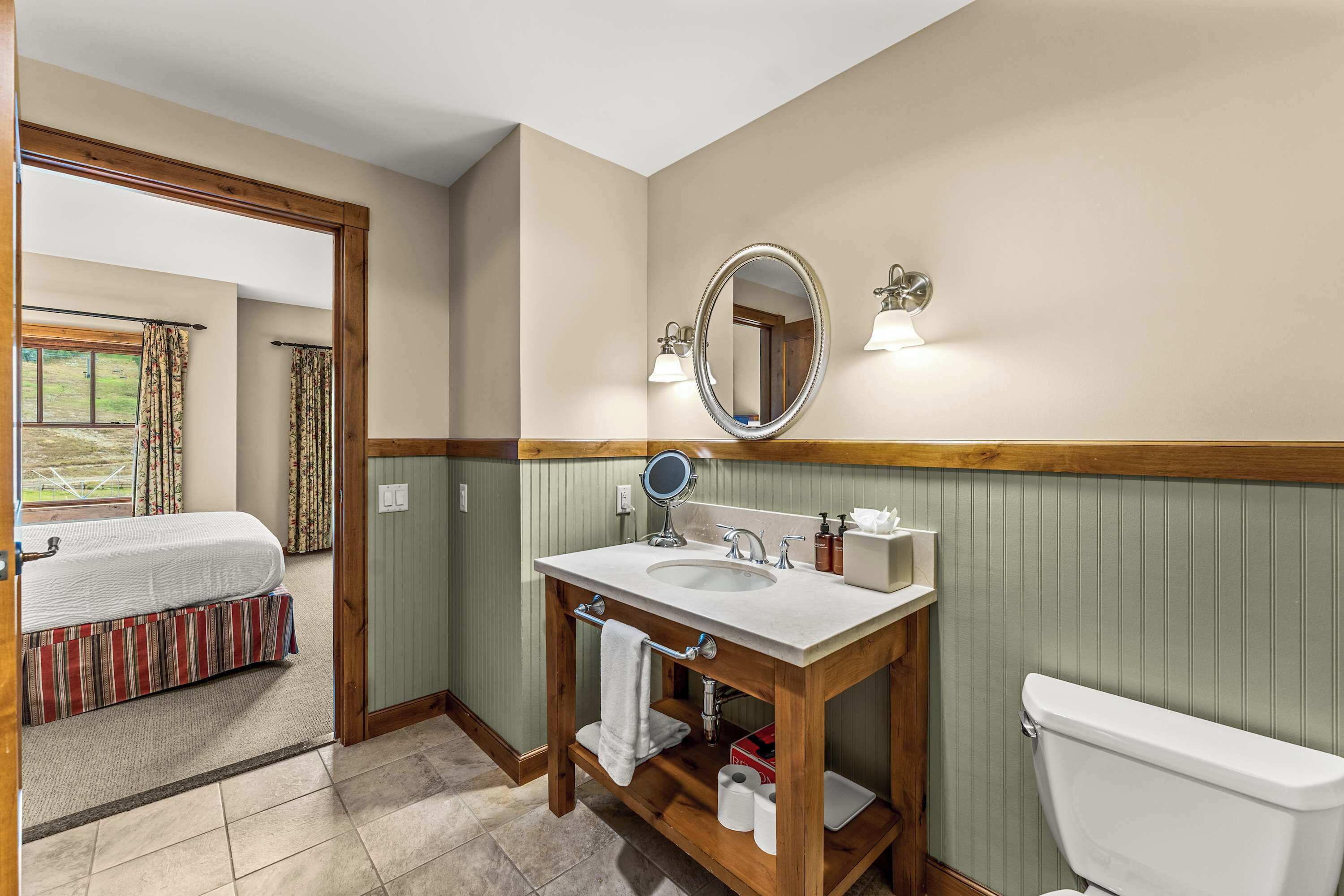 A bathroom with a wooden vanity, a double sink, a toilet, and a bed in the adjacent room.