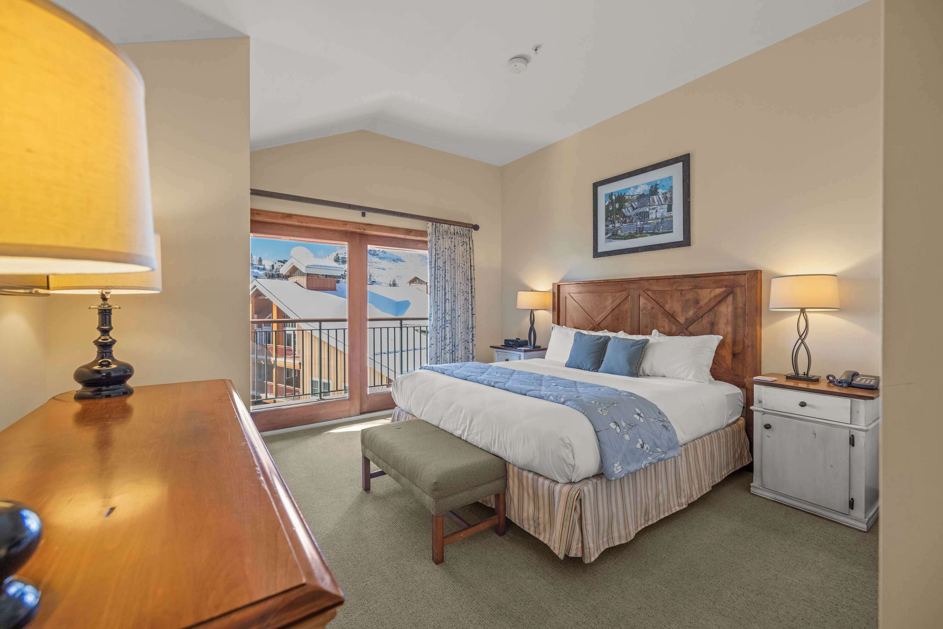 A hotel room with a large bed, a wooden nightstand, a desk, and a balcony with a view of a snowy landscape.