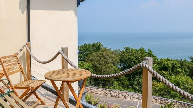 Ein Balkon mit Geländer aus Seil, Holzstühlen und einem Tisch mit Blick auf eine malerische Landschaft aus Meer und Grün.