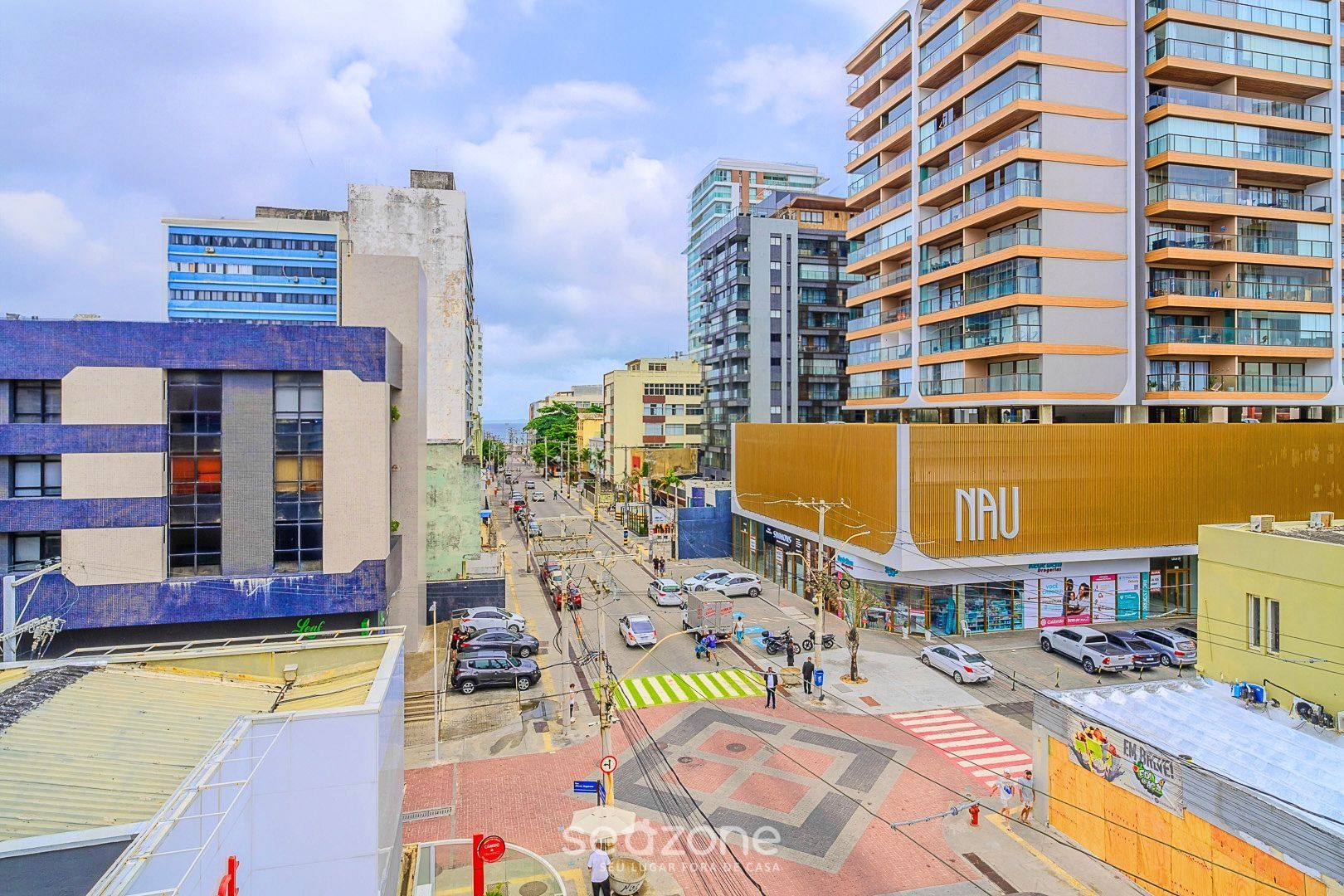 Uma rua urbana com edifícios modernos, um edifício amarelo de destaque com a sinalização 'NAU' e uma faixa de pedestres.