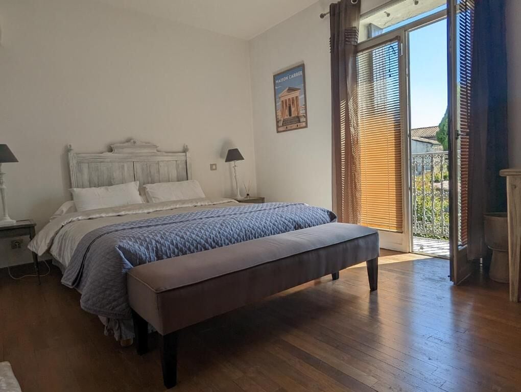 Une chambre à coucher avec un lit en bois, un banc, une table de chevet, une lampe et une vue sur l’extérieur par une porte munie de stores.