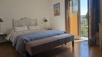 Une chambre à coucher avec un lit en bois, un banc, une table de chevet, une lampe et une vue sur l’extérieur par une porte munie de stores.