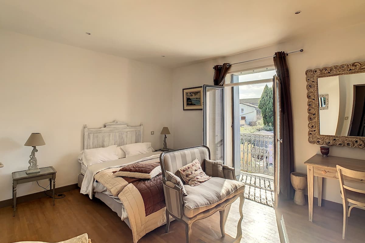 A bedroom with a bed, two chairs, a table, a mirror, and a window with a view of the outside.