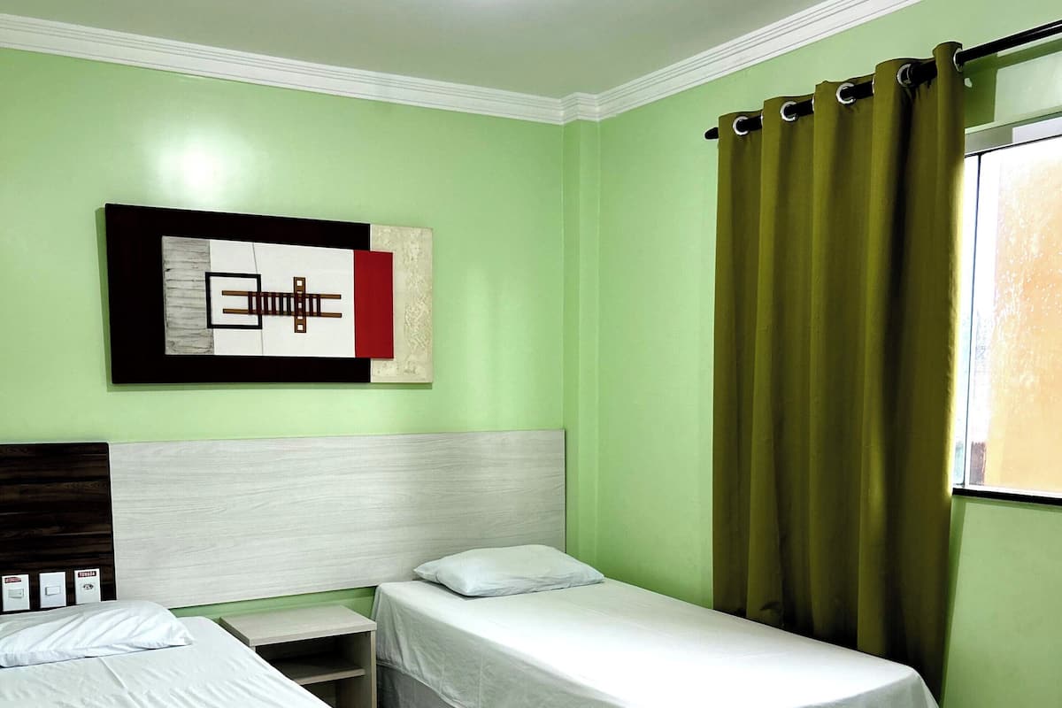 Una habitación con dos camas individuales, una mesita de noche, un aplique de pared y una ventana con cortinas.