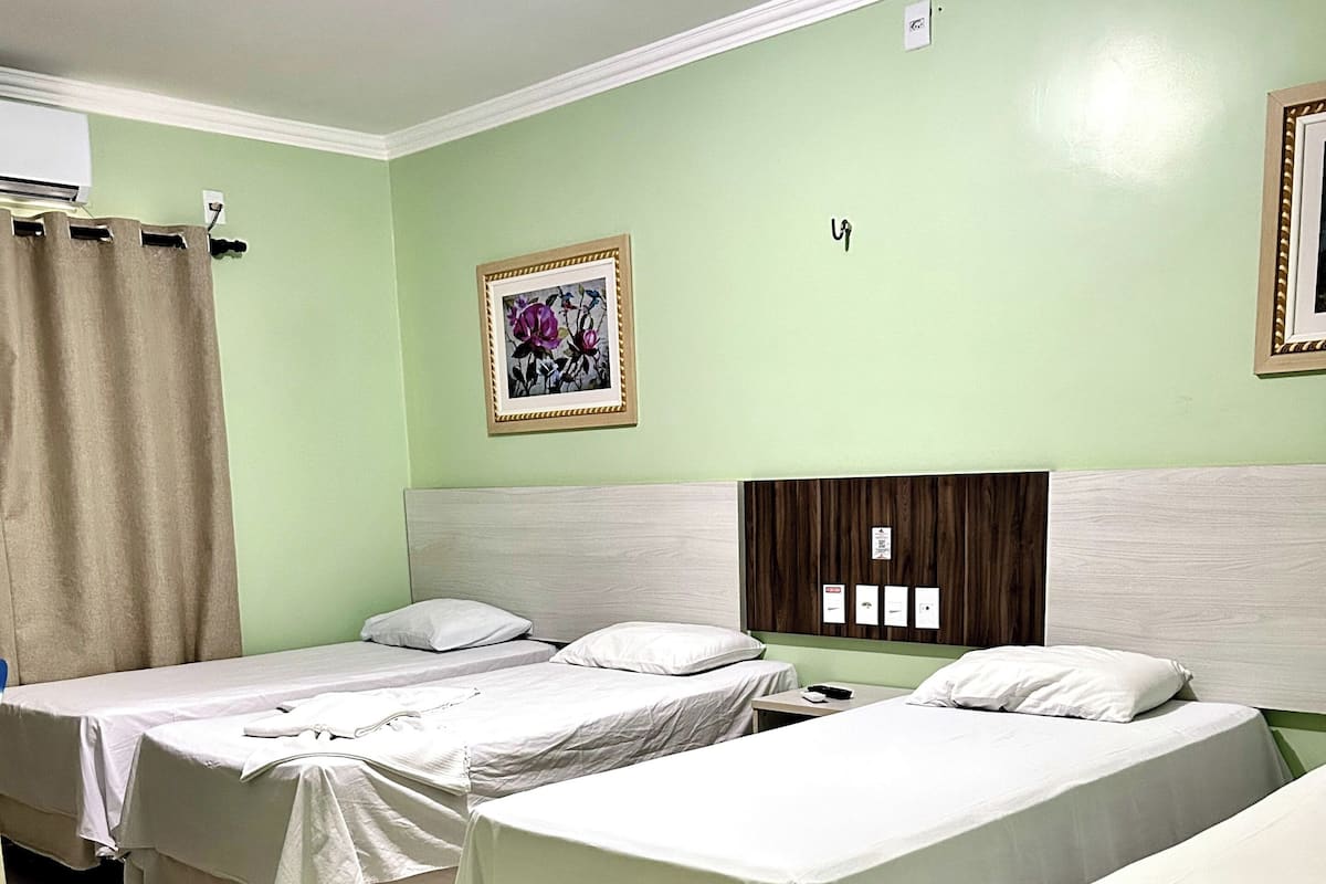Habitación de hotel con tres camas individuales, cabecera de madera, aire acondicionado montado en la pared y una obra de arte floral enmarcada.