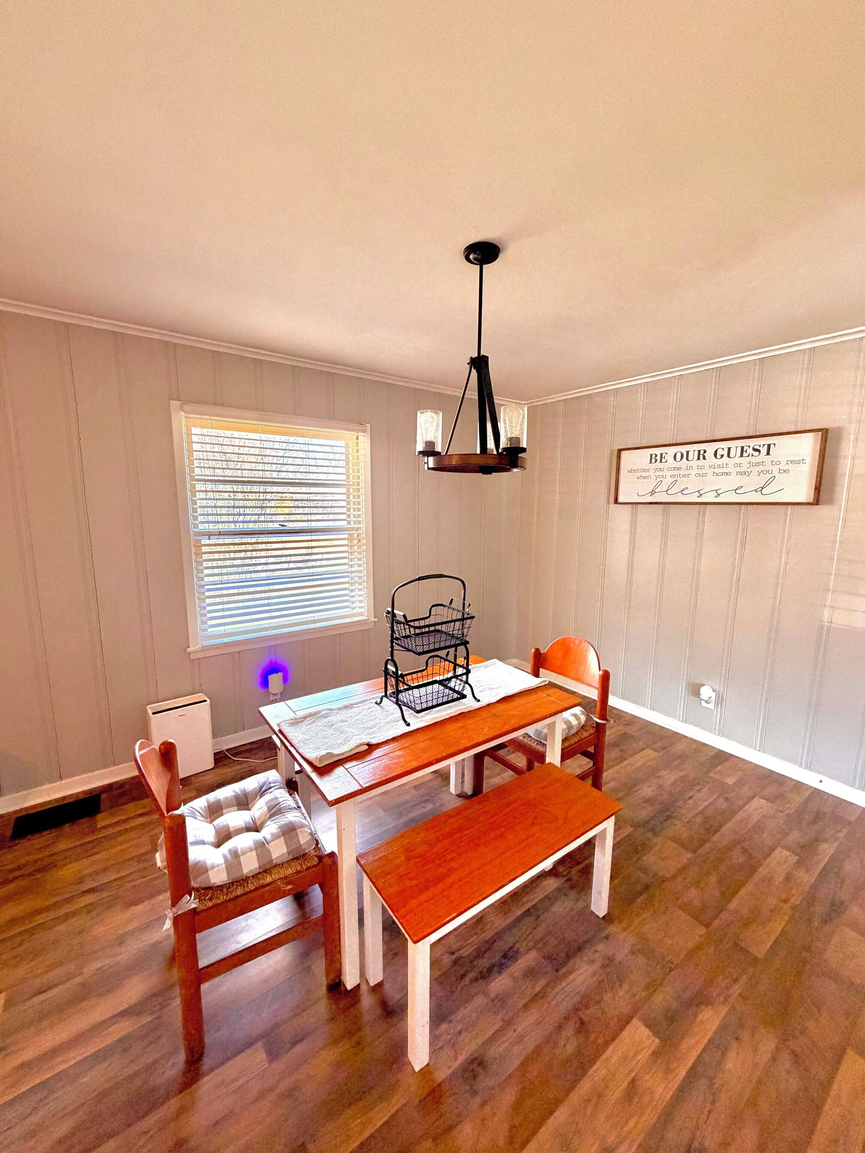 A dining area with a wooden table, chairs, a bench, a chandelier, a window with blinds, and a wall-mounted sign.