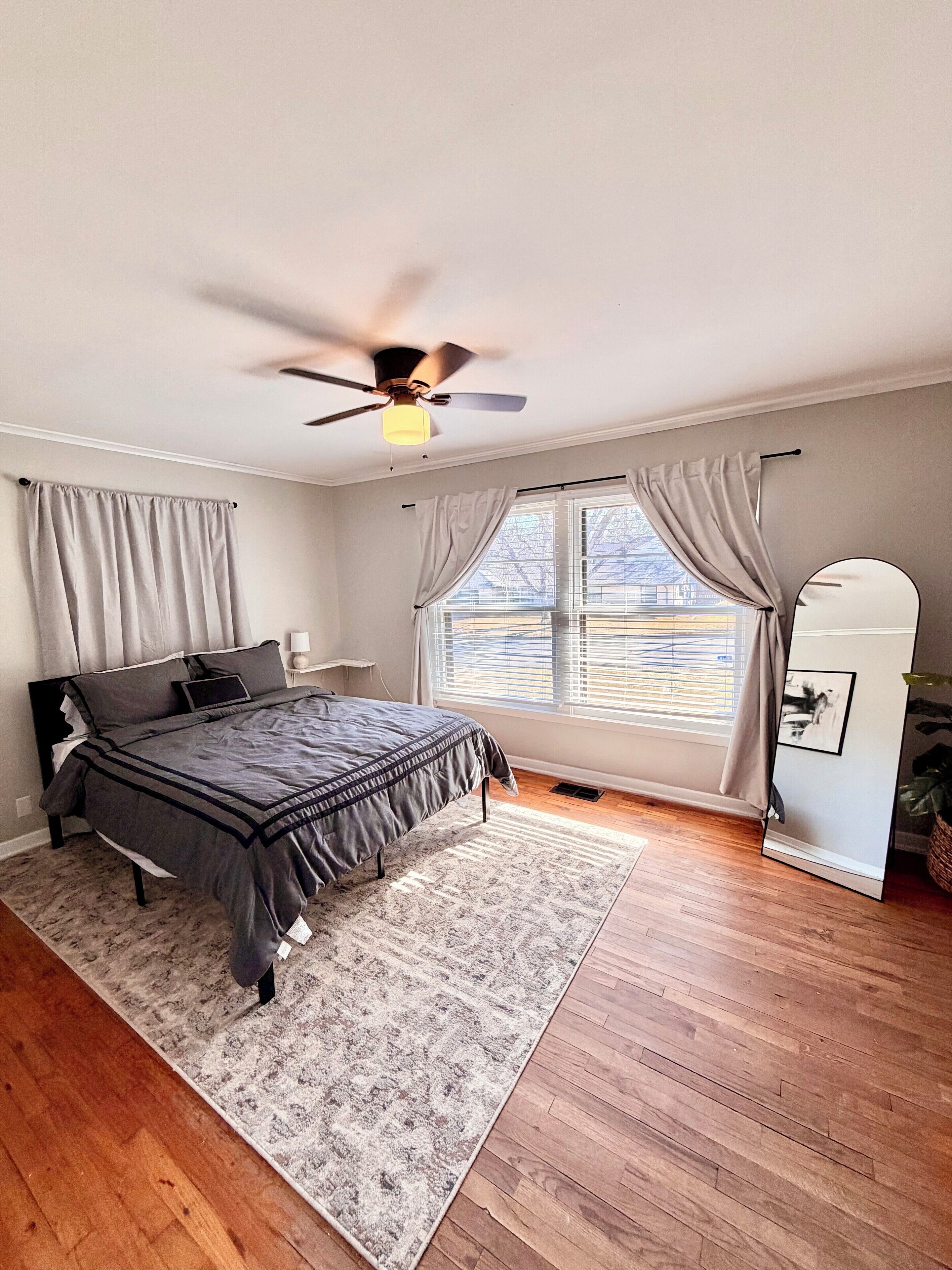 Un dormitorio con cama, ventilador de techo, ventana con cortinas, mesita de noche y espejo.