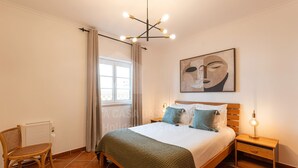 Ein Schlafzimmer mit einem Bett, einem Nachttisch, einem Sessel, einem Fenster mit Vorhängen und einer Deckenleuchte.