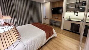 Moderni hotellihuone, jossa on sänky, yöpöytä, lamppu sekä pieni keittiöalue, jossa on televisio ja kaappeja.