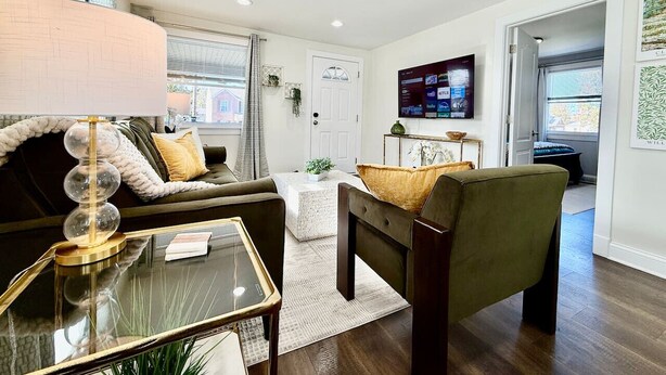 A modern living room with a sofa, armchair, and coffee table. There is a television mounted on the wall and a door leading to another room. - Cute & Cozy House in Quiet Blue Ash Neighborhood (Blue Ash)