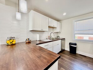 A kitchen with white cabinets, a wooden countertop, a window with blinds, and a trash can. - Cute & Cozy House in Quiet Blue Ash Neighborhood (Blue Ash)