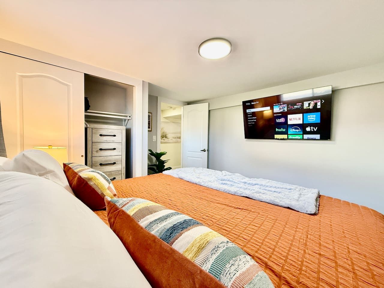A hotel room with a bed, a TV mounted on the wall displaying various streaming services, a bedside table with a lamp, and a wardrobe.