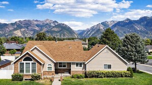A two-story house with a brick facade and a sloped roof, surrounded by a well-maintained lawn and a mountain backdrop. - Snowbird & Alta Ski Retreat + Walk to Ski Bus + High Ceilings + Newly Remodeled (Sandy)