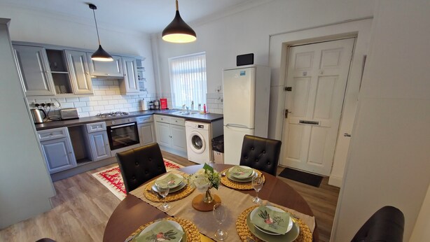 A dining area with a table set for four, a kitchen in the background, and a door to the right. - Ushaw Hilltop View- Comfortable 2-Bed Home Near Durham University & City (Ushaw Moor)