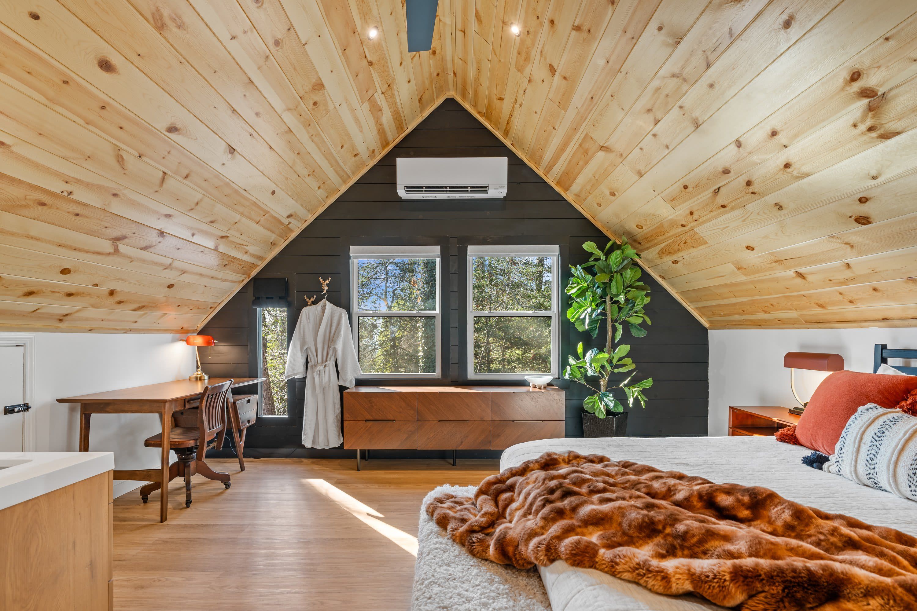 A cozy attic bedroom with wooden slats on the ceiling, a large window, a bed, a desk, and a chair.