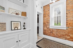 A hallway with a white wall, a brick wall, and a window with blinds. There are shelves with framed pictures and a door leading to another room. - Highlands Retreat w/Hot Tub, Parking & Large Fenced Back Yard (Louisville)
