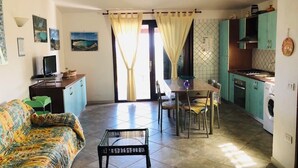 Una sala de estar con sofá, una mesa de comedor y sillas, una cocina equipada con lavadora, y un televisor.