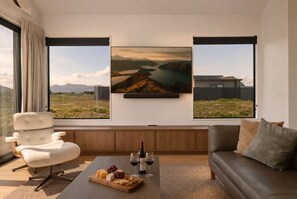 A modern living room with a large window, a white sofa, a leather couch, a wooden coffee table with a cheese board, wine glasses, and two paintings of landscapes. - Modern Wanaka Escape with Panoramic Mt Roy Views (Wānaka)