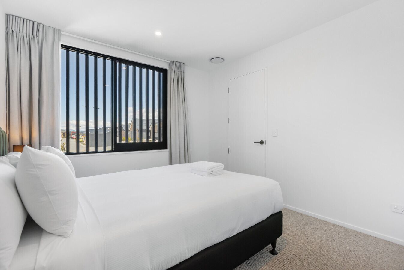 A neatly made bed with white linens and pillows, a window with vertical blinds, and a door to the right.