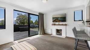 A modern living room with a fireplace, a large window, and a sliding glass door to an outdoor deck. - Central Wanaka Escape | Lake Views | Walk to Town | Deck & BBQ (Wānaka)