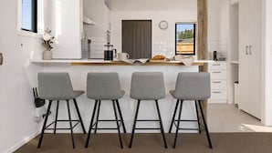 A modern kitchen with white cabinetry, a central island with bar stools, and a large wooden beam. - Central Wanaka Escape | Lake Views | Walk to Town | Deck & BBQ (Wānaka)