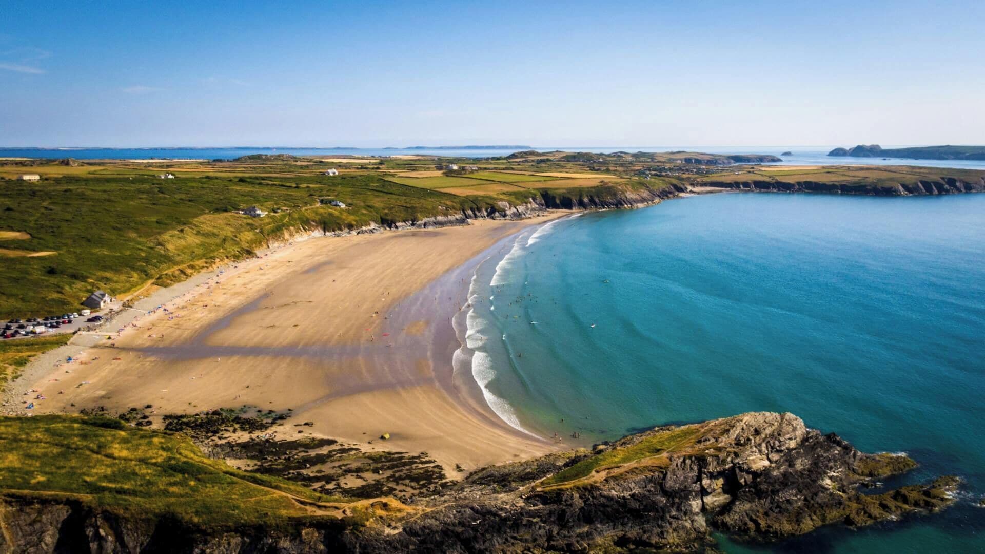 A coastal landscape with a sandy beach, clear blue waters, and rocky cliffs.
