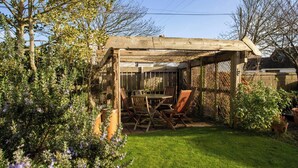 Eine hölzerne Pergola mit Tisch und Stühlen, umgeben von einem Garten mit verschiedenen Pflanzen und einer Rasenfläche.