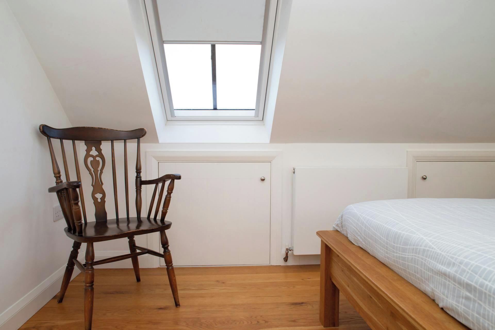 A bedroom with a sloped ceiling, a wooden chair, a bed with a white bedspread, and a white radiator.