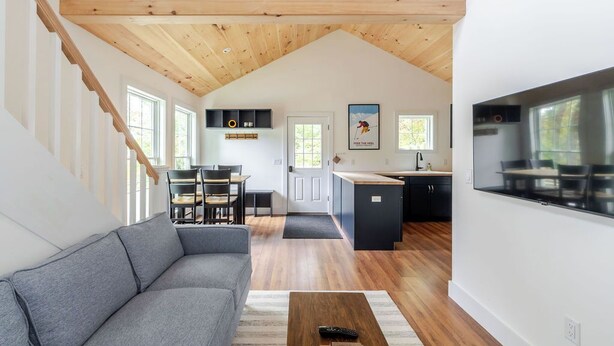 Un intérieur moderne avec un plafond de style industriel, un canapé gris, une table basse en bois et un coin cuisine doté d’une grande télévision.