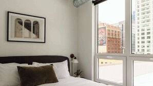 A hotel room with a large bed, a framed picture on the wall, a nightstand with a lamp, and a view of the city through large windows. - Luxury Penthouse Loft w/Terrace  504 | River North (Chicago)