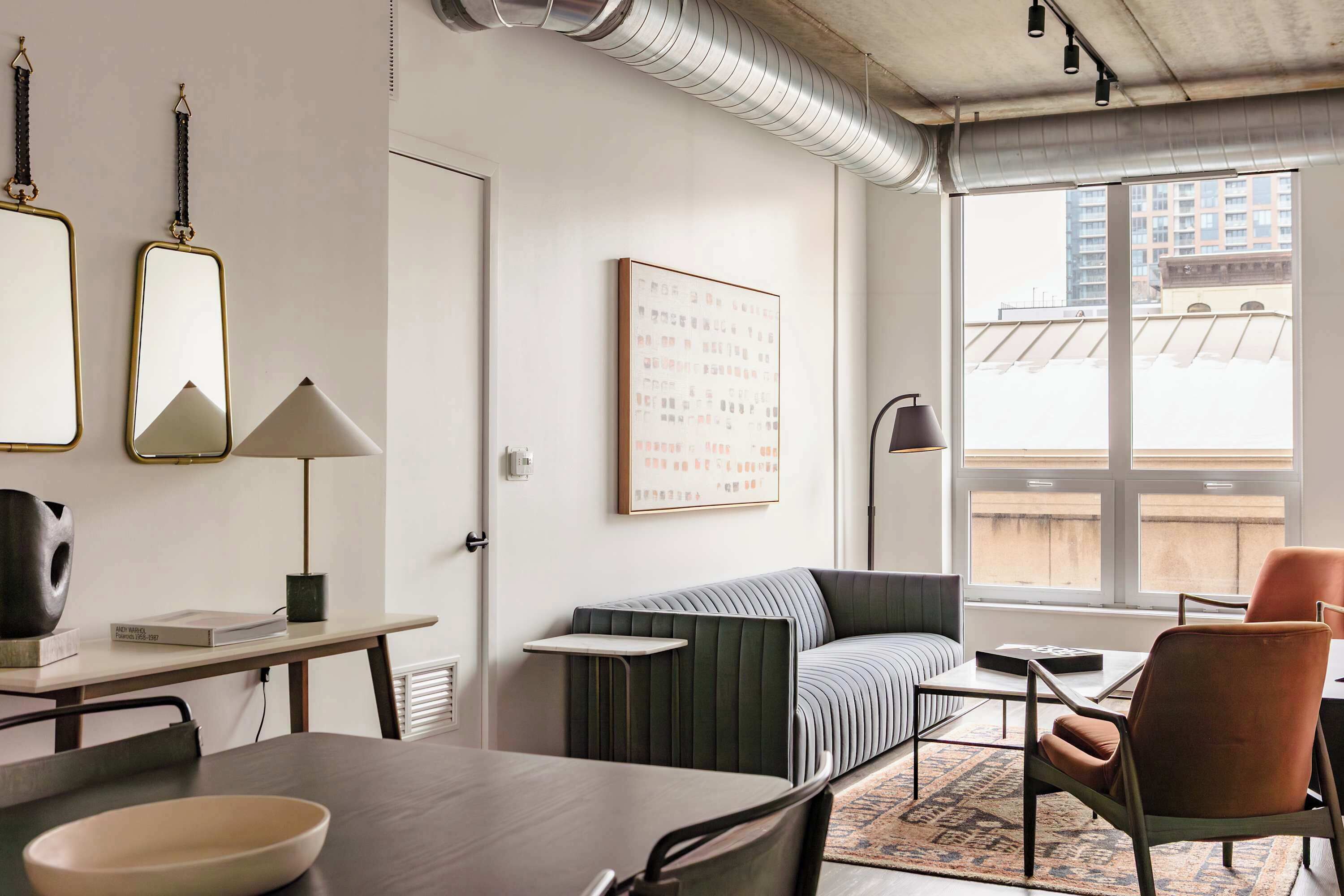 A modern living room with a sofa, a dining table, and a coffee table. There is a large window with a view of buildings outside.