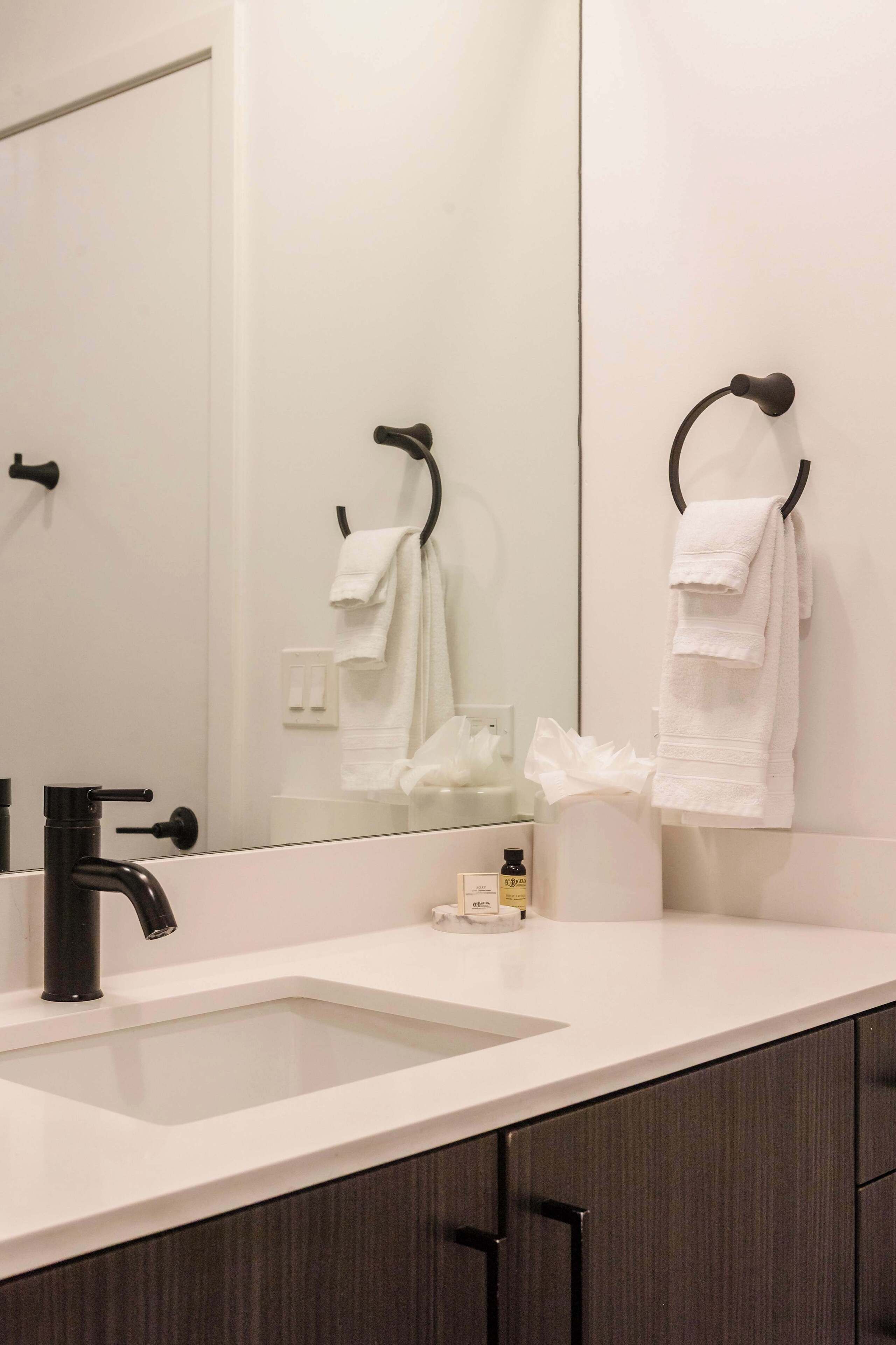 A modern bathroom with a white sink, black faucet, and a large mirror. There are white towels hanging on hooks and a tissue box on the counter.