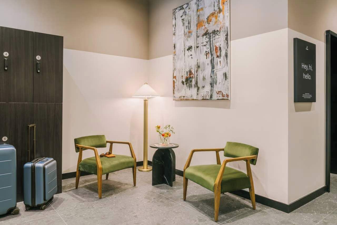 A hotel lobby with two green chairs, a table with a vase, a lamp, and a large abstract painting on the wall.